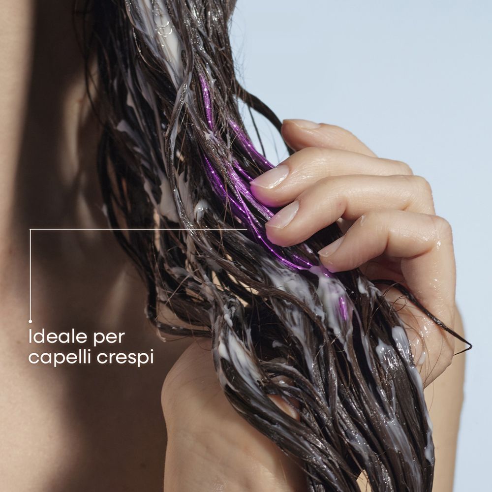 Mano che applica balsamo bianco su capelli scuri e bagnati con riflessi viola. Ideale per capelli crespi.