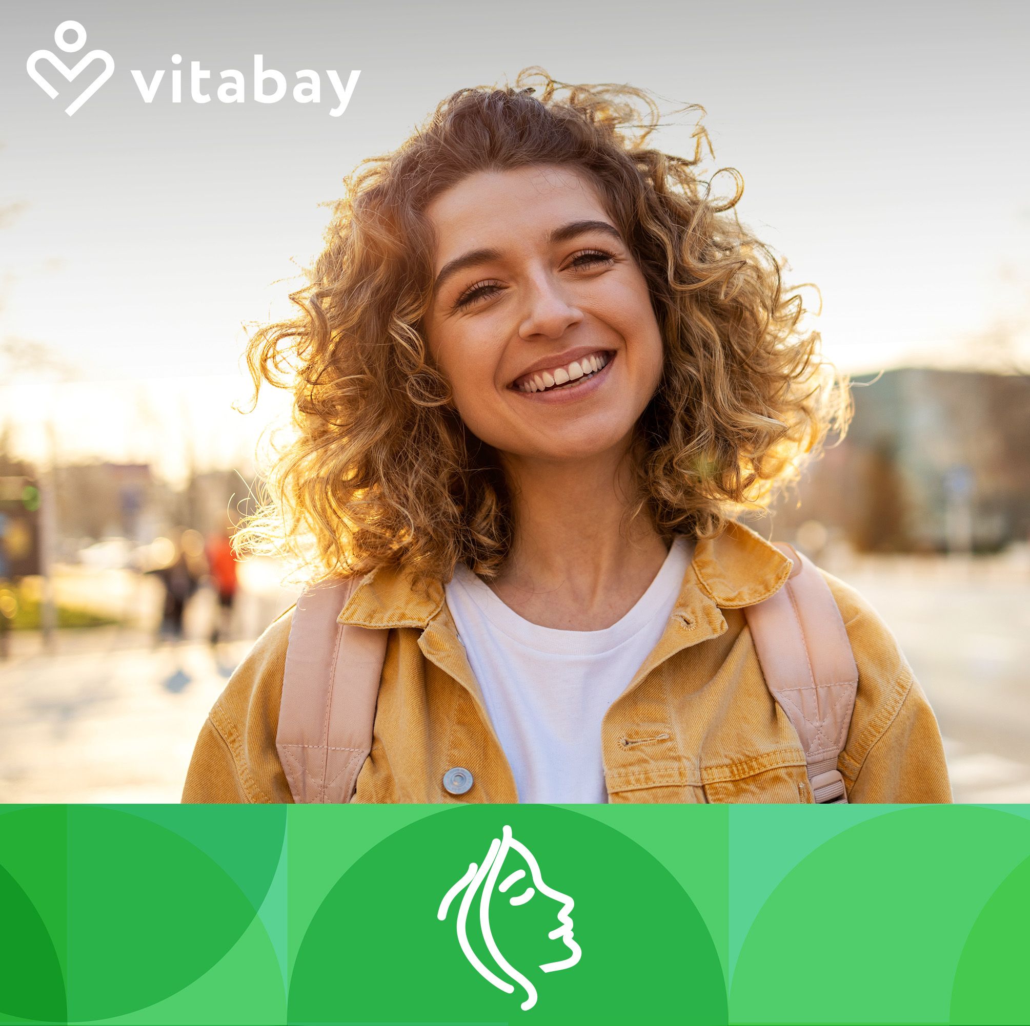 Una donna sorridente con i capelli ricci e la luce del sole sullo sfondo. Indossa una giacca gialla e uno zaino.