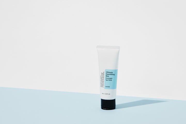 Tubo bianco con tappo nero. Scritta: Ultimate Nourishing Rice Overnight Spa Mask. Marchio: COSRX. Su sfondo blu.