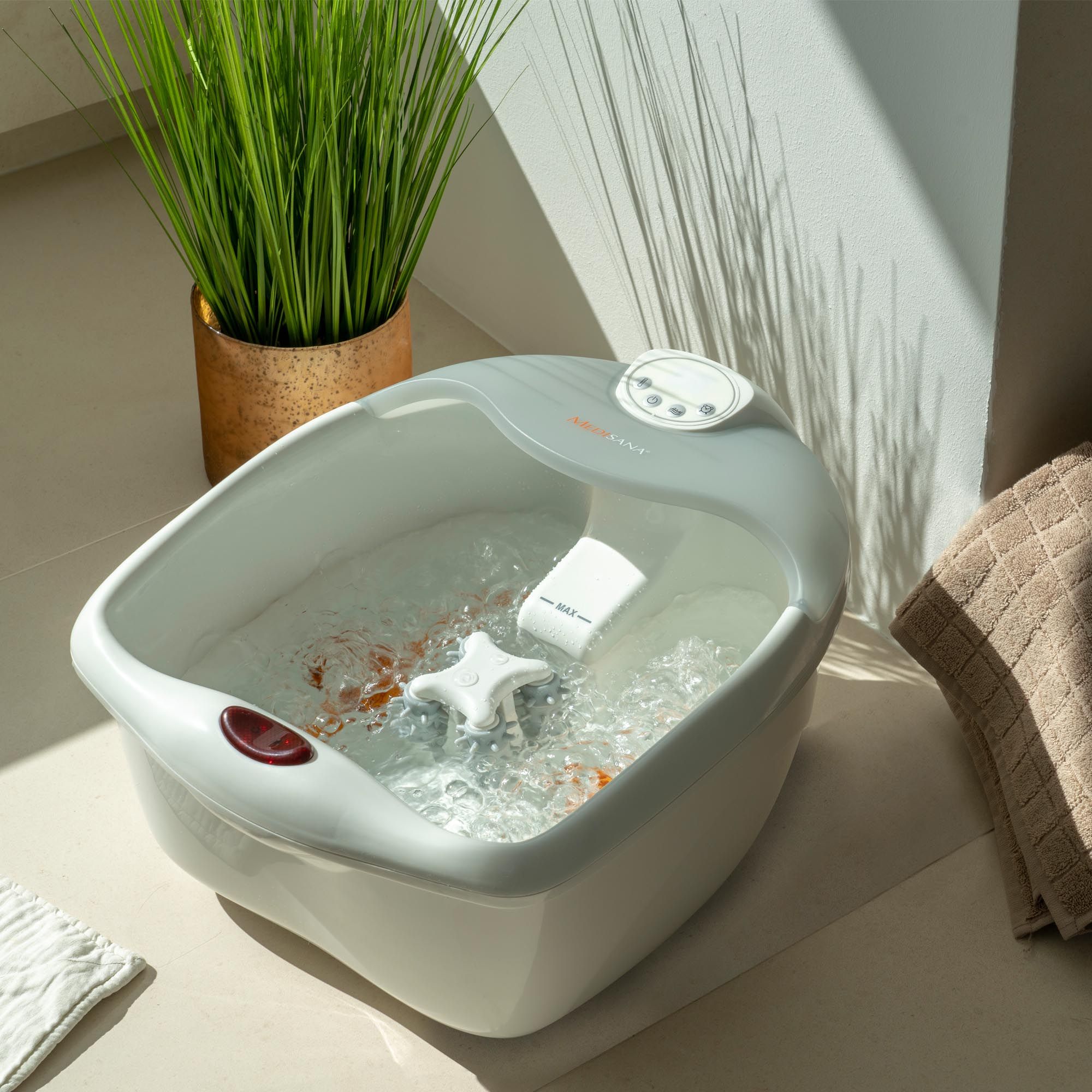 Idromassaggio per piedi con acqua, circondato da piante. Bianco-grigio, pulsante rosso, pannello di controllo. Acqua con bolle.
