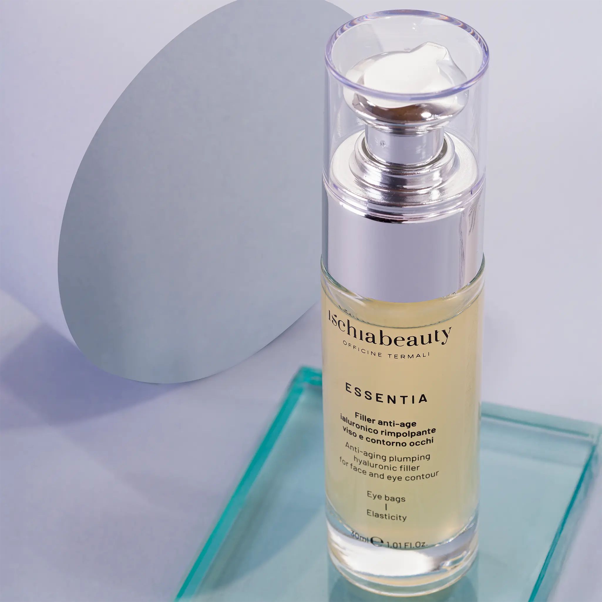 Flacone in vetro con pompa argentata. Scritte: ischiabeauty, ESSENTIA, Filler anti-age, borse occhi, elasticità. Su lastra di vetro.