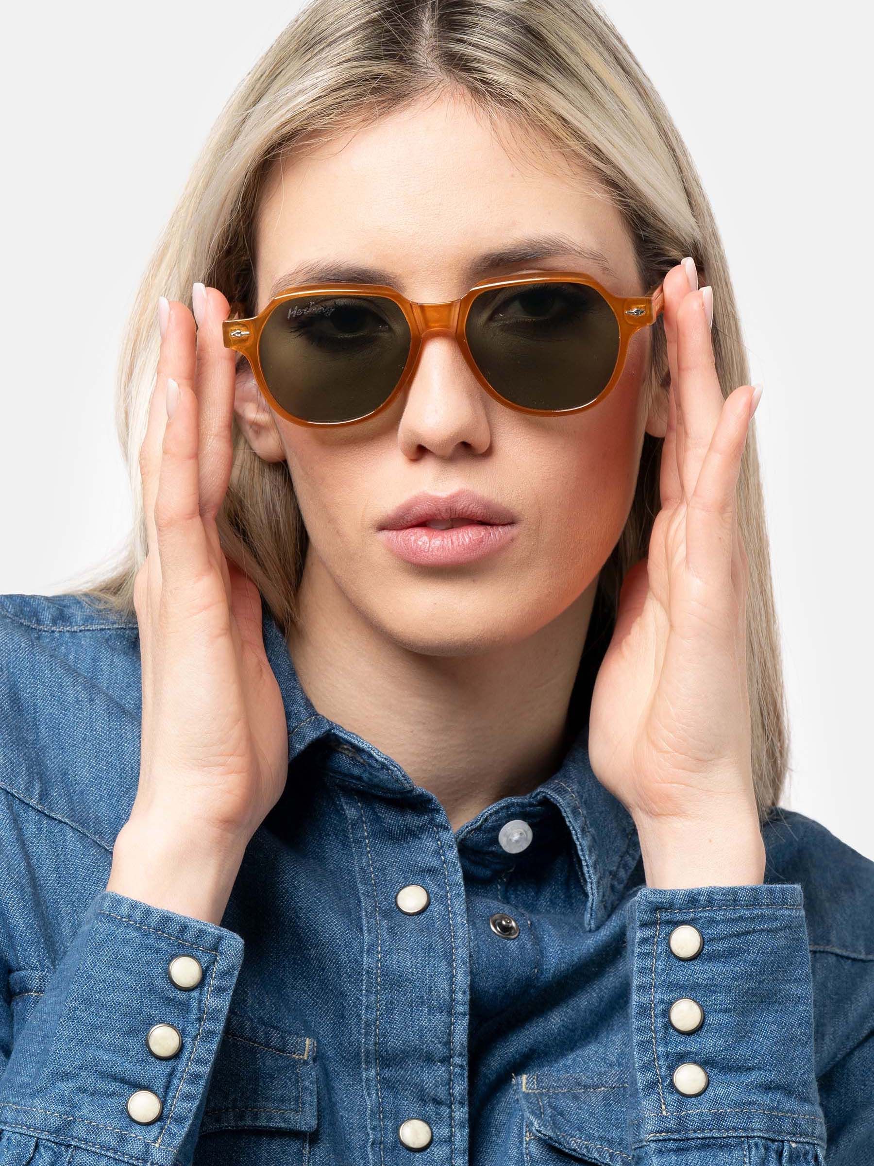 Donna che tiene occhiali da sole color ambra con lenti verdi. Forma esagonale. Marchio Herling visibile.