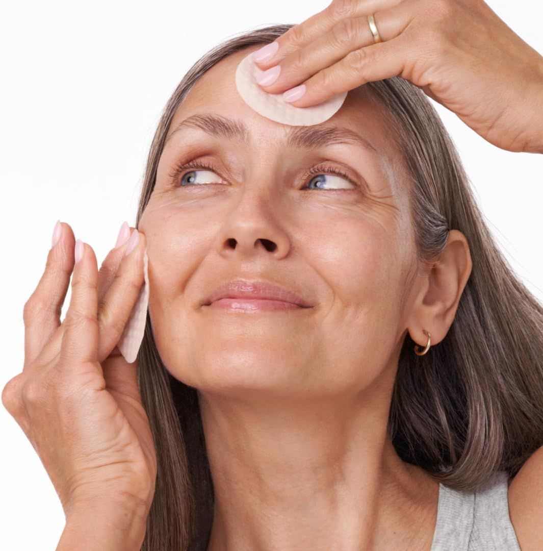 Donna con dischetti per il viso. Mani che tengono i dischetti sulla fronte e sulla guancia. Capelli grigi, top grigio. Guarda in alto.