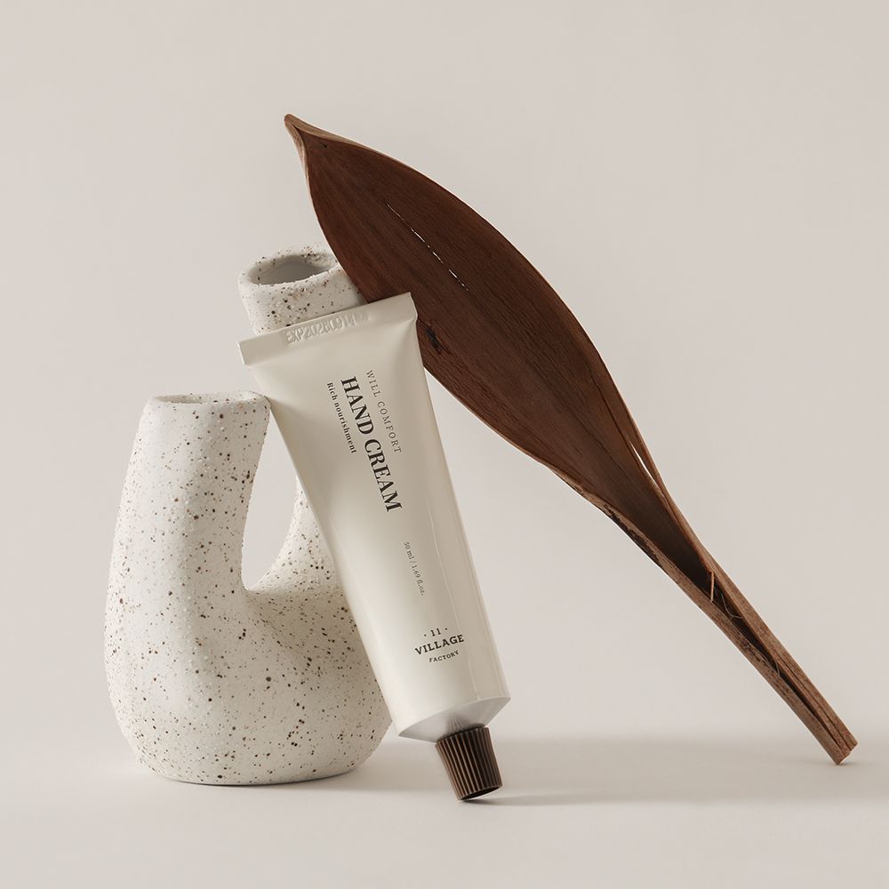 Tubo di crema, accanto a un vaso e una foglia. Scritta: HAND CREAM, WILL COMFORT, VILLAGE FACTORY. Tappo marrone.