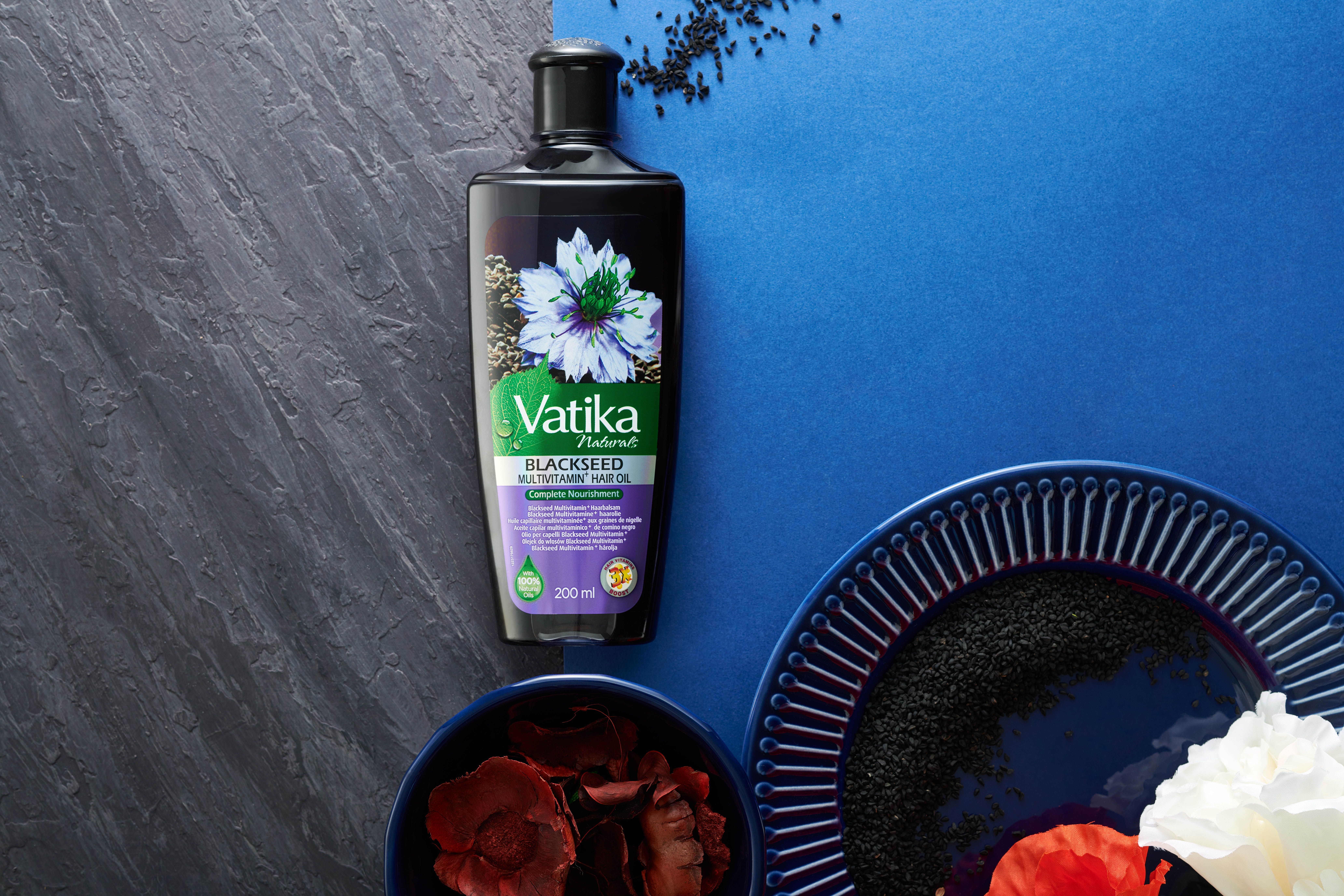 Olio per capelli Vatika Naturals Schwarzkümmel su sfondo blu. Accanto, ciotole con ingredienti e un piatto.