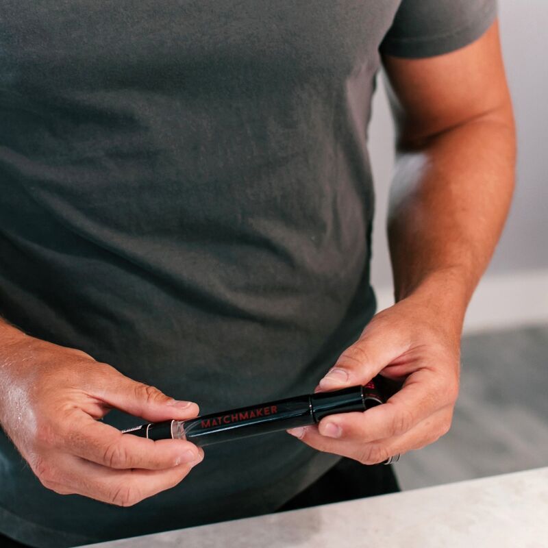 Uomo che tiene un flacone di profumo nero. Scritta rossa "MATCHMAKER" sul flacone. L'uomo indossa una maglietta grigia.