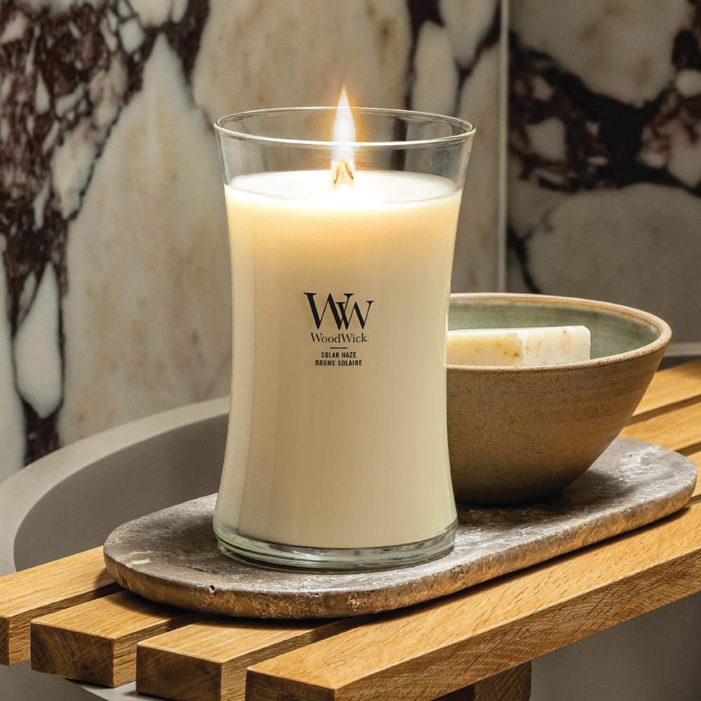Candela accesa in vetro. Marchio WoodWick visibile. Accanto a sapone e ciotola su vassoio di legno.