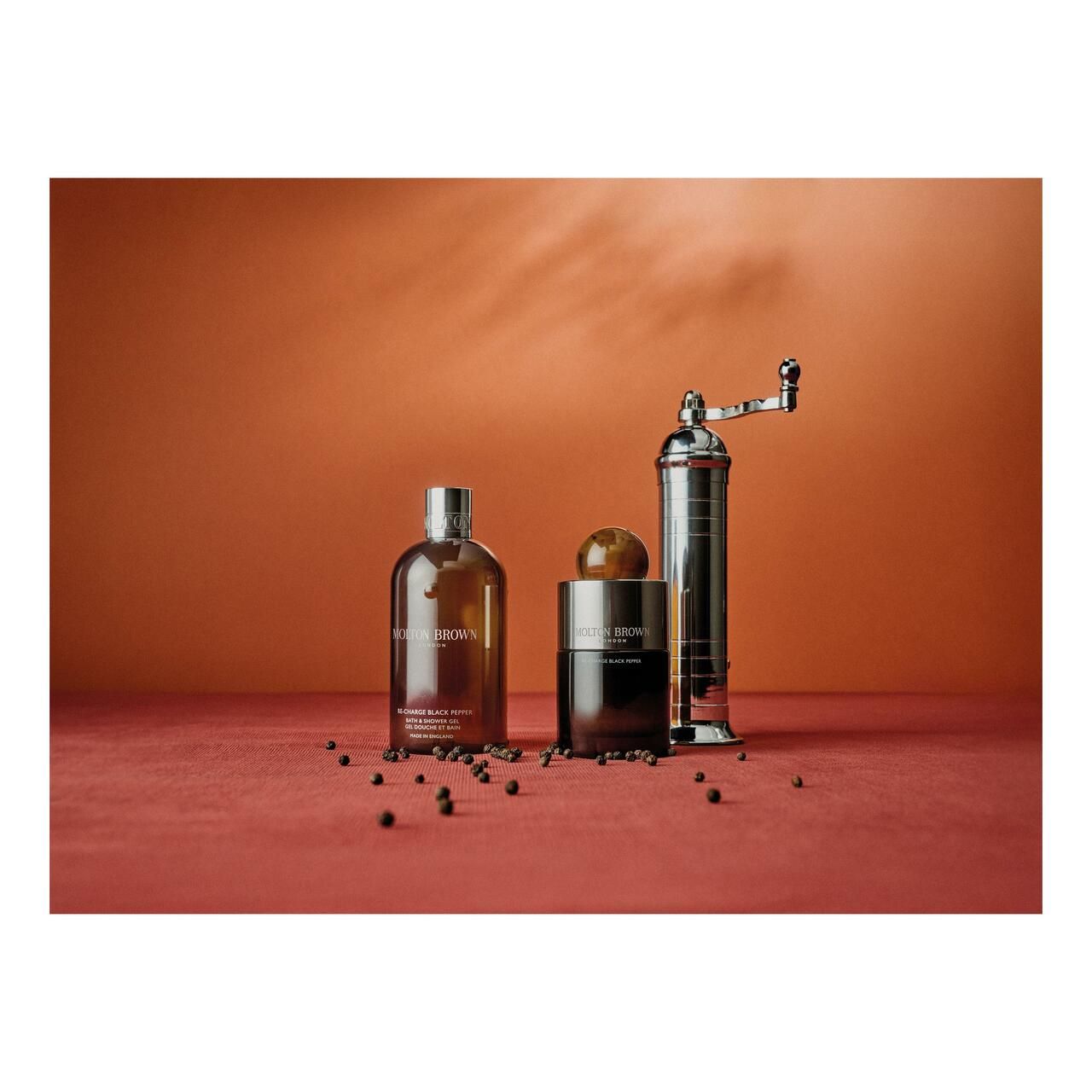 Tre prodotti Molton Brown su sfondo rosso. Una bottiglia, un vasetto e un macinapepe.