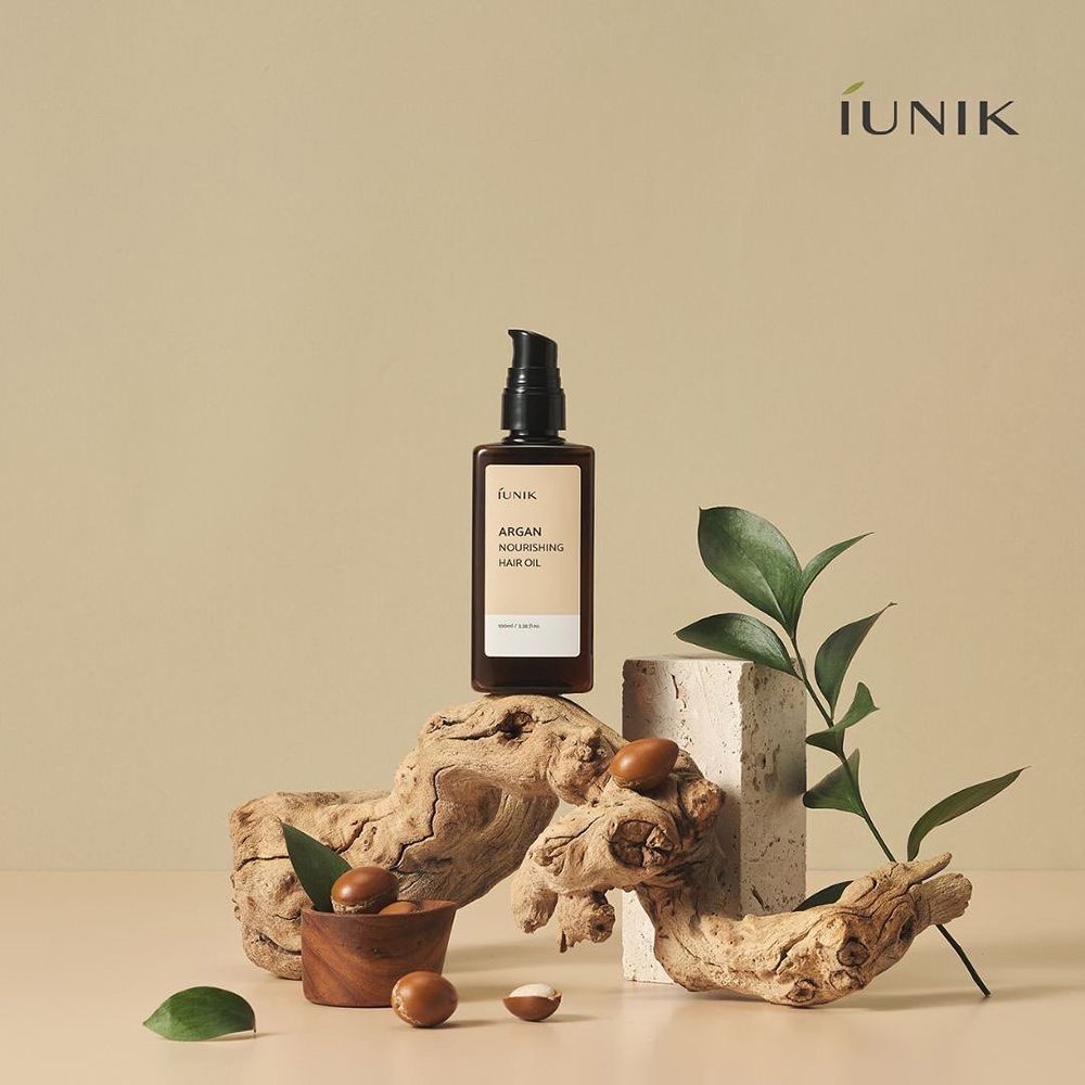 Flacone marrone con erogatore nero. Posizionato su legno e pietra. Noci di argan e foglie. Scritta: IUNIK, Argan Nourishing Hair Oil.