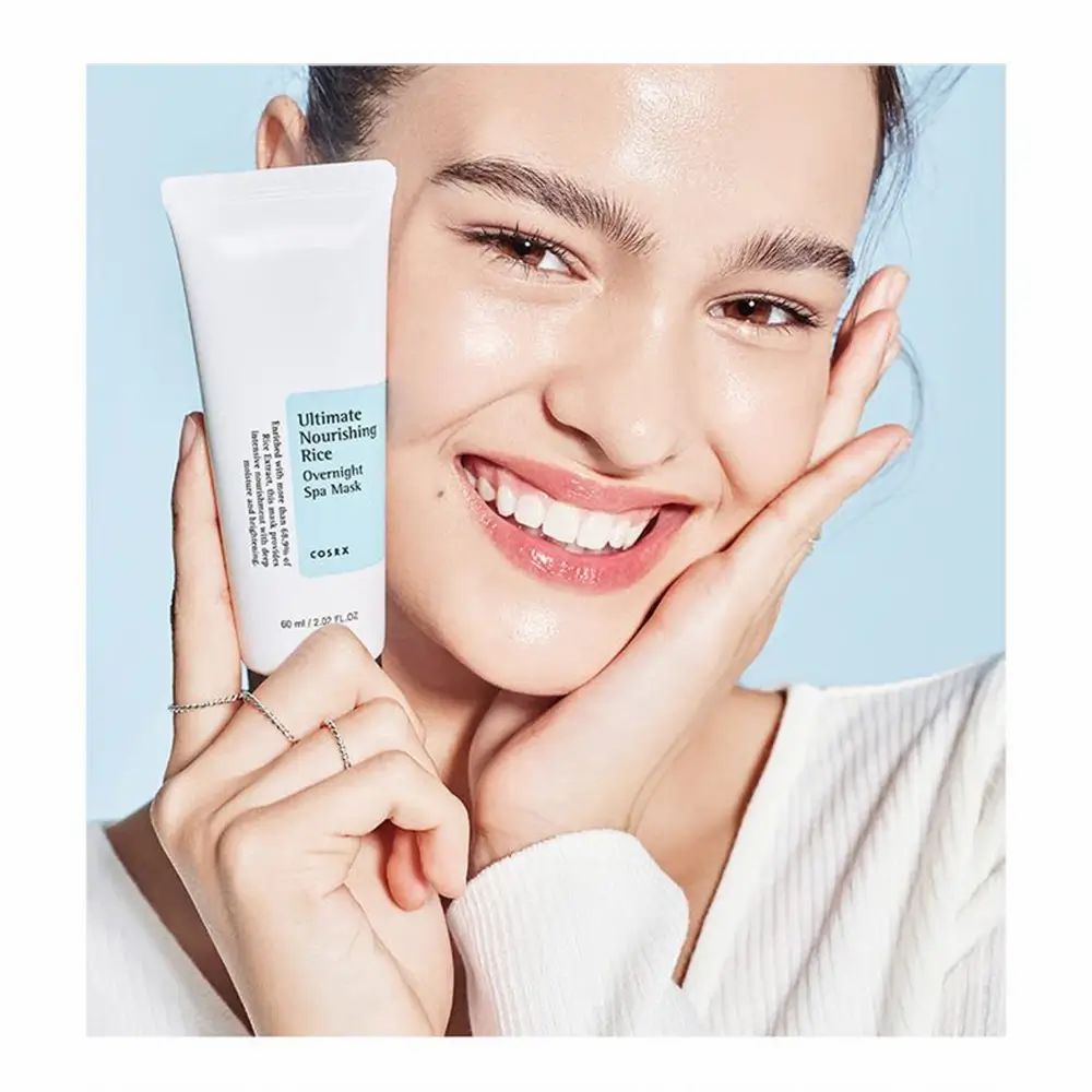 Donna che tiene un tubo con scritta: Ultimate Nourishing Rice Overnight Spa Mask. Marchio: COSRX. Su sfondo blu.
