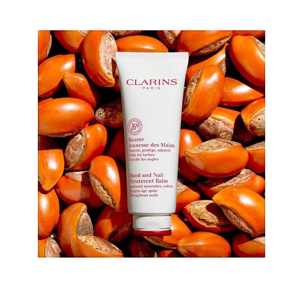 Tubo bianco con scritta Clarins, Balsamo per il trattamento delle mani e delle unghie. Circondato da semi marroni. Contiene il 26% di ingredienti.