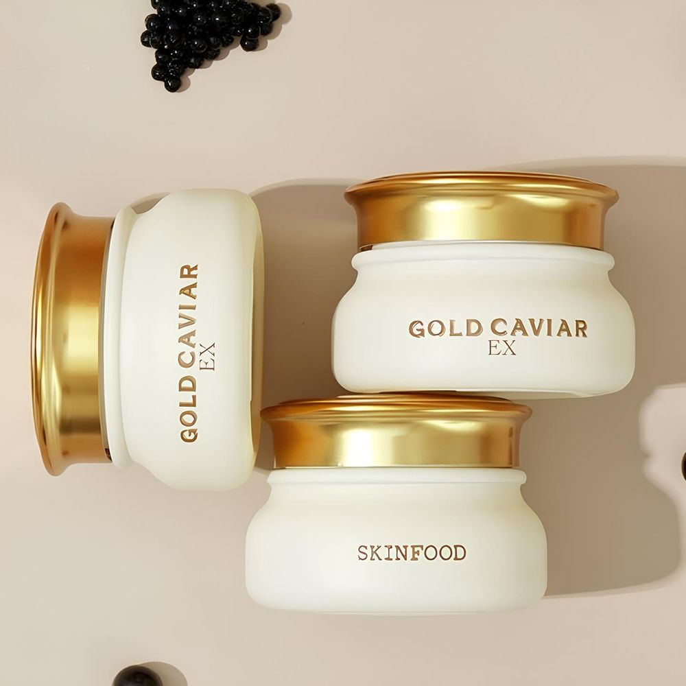 Tre vasetti di crema con coperchi dorati. Scritta: GOLD CAVIAR EX e SKINFOOD. Su sfondo rosa chiaro.