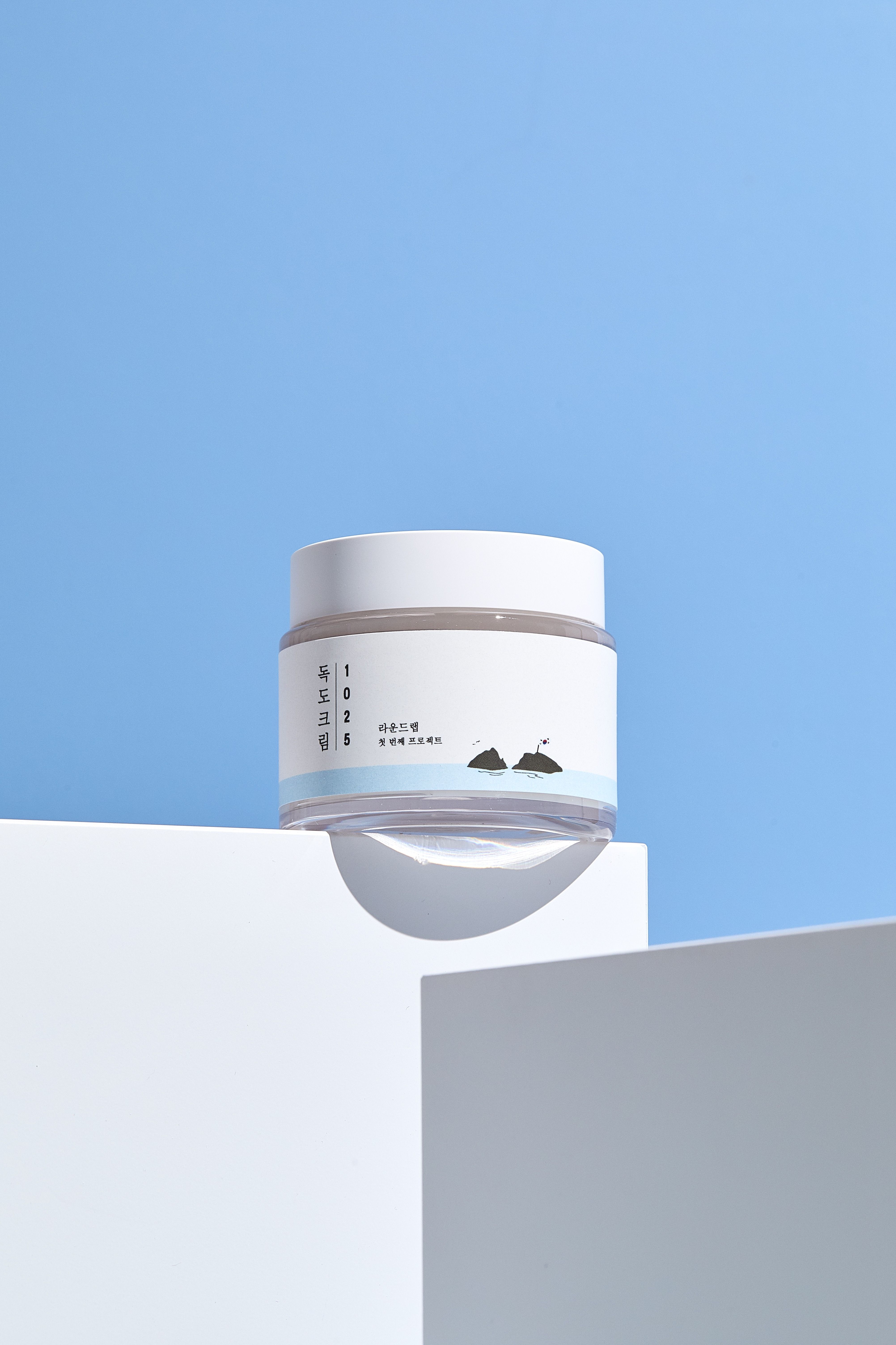 Vasetto di crema bianco su piedistallo bianco. Scritta coreana e numeri. Sfondo blu.