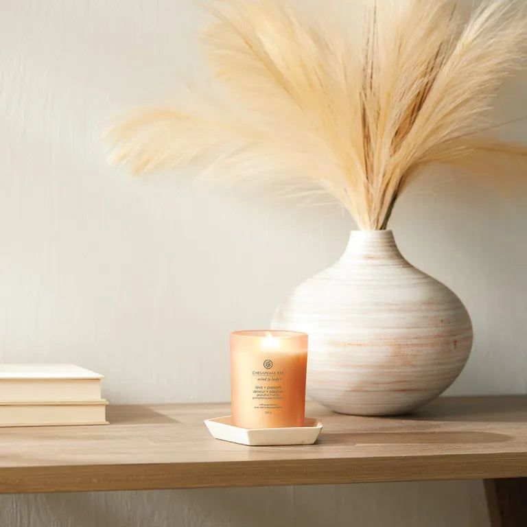 Candela in contenitore di vetro su sottobicchiere bianco. Accanto a vaso con erba della pampa. Su superficie in legno.