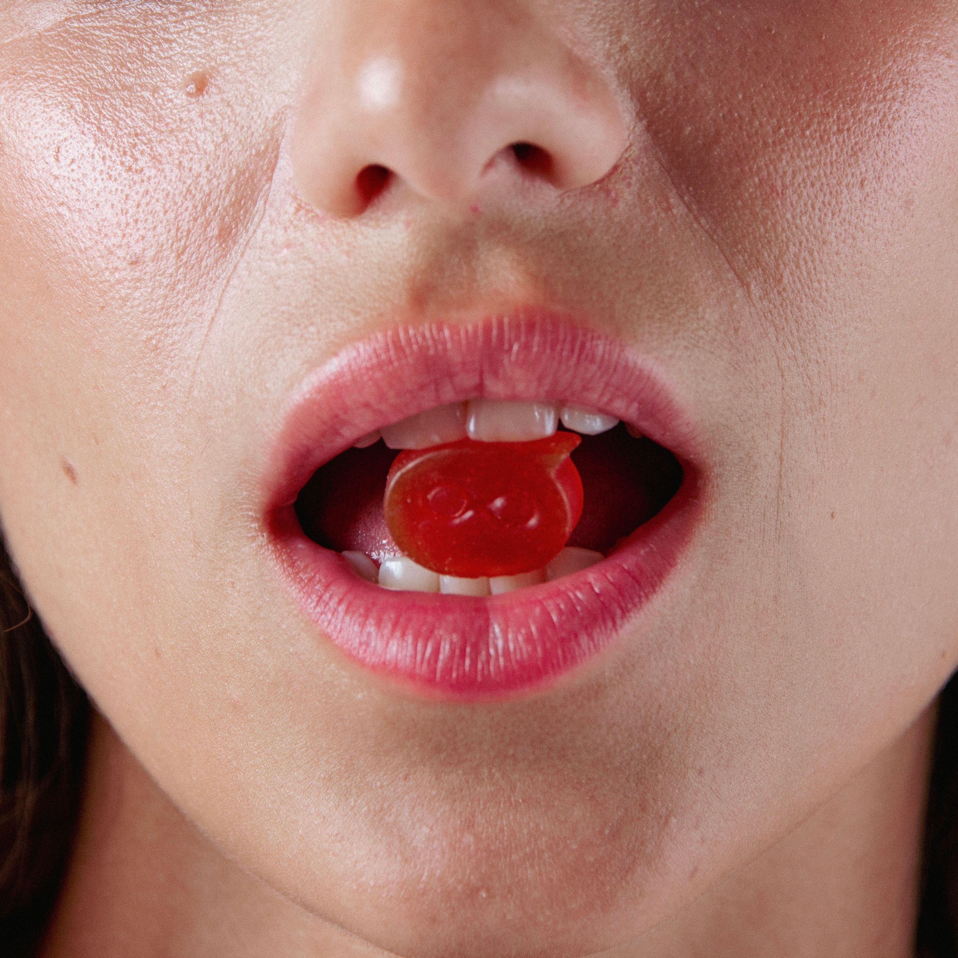 Primo piano di una bocca con una caramella gommosa rossa. Le labbra sono rosa e aperte.