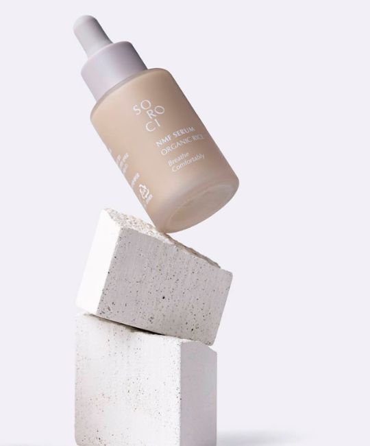 Flacone di siero su blocchi di pietra. Tappo bianco, scritta. Flacone beige. Scritta: NMF Serum Organic Rice.