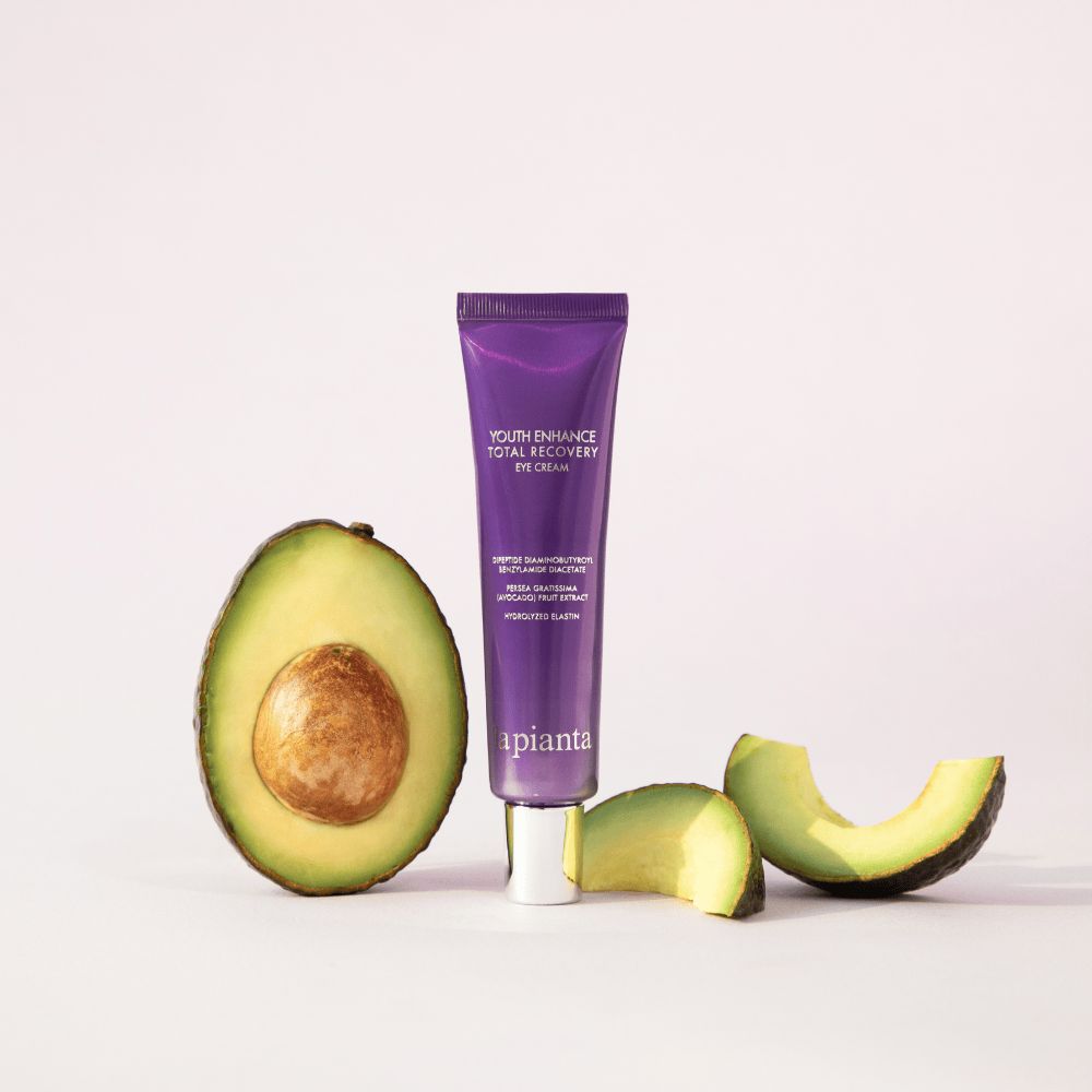 Tubo viola con tappo bianco. Metà di avocado accanto. Scritta: YOUTH ENHANCE TOTAL RECOVERY EYE CREAM, la pianta.
