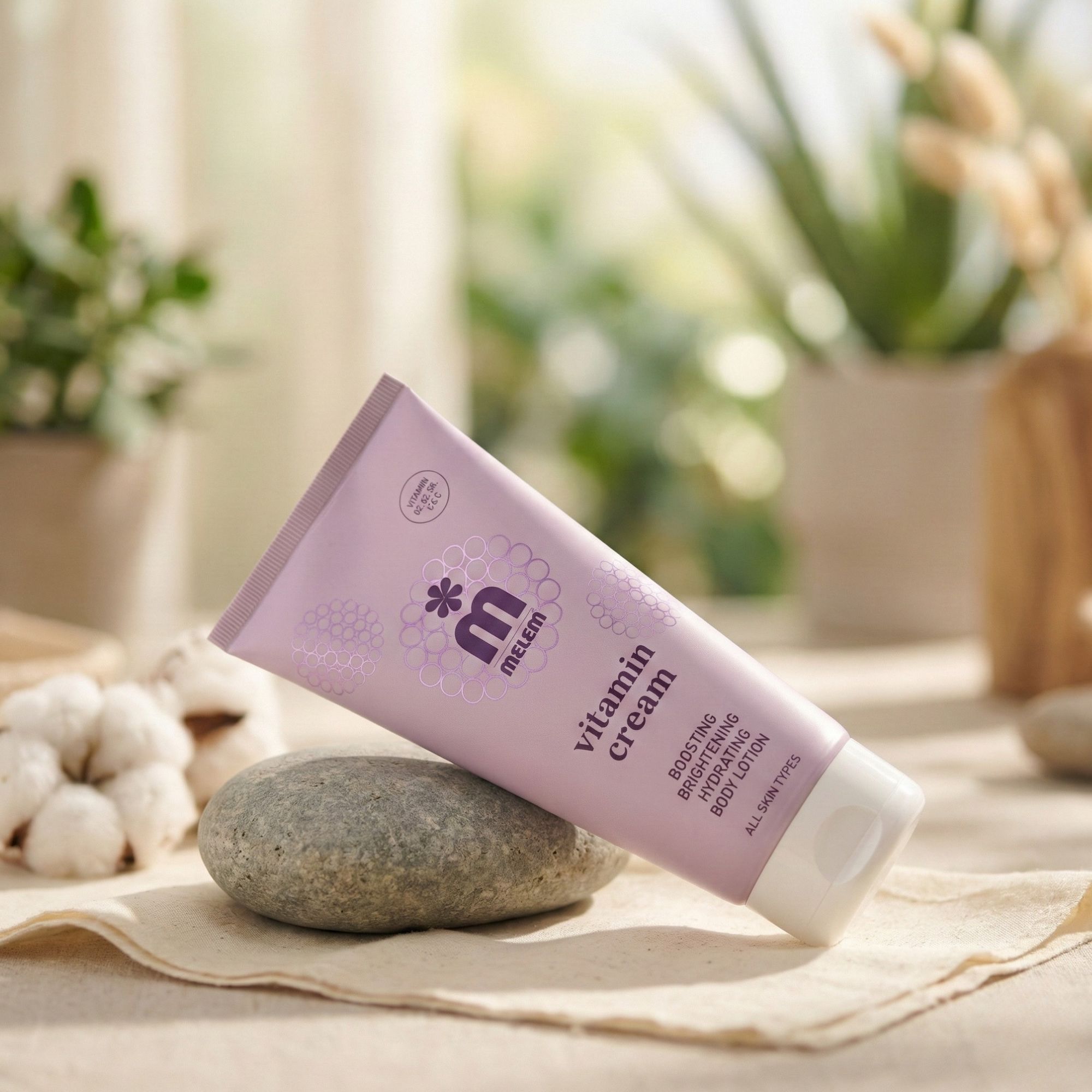 Tubo rosa con tappo bianco. Scritta: Melem Vitamin Cream. Appoggiato su una pietra. Sullo sfondo: piante e cotone.