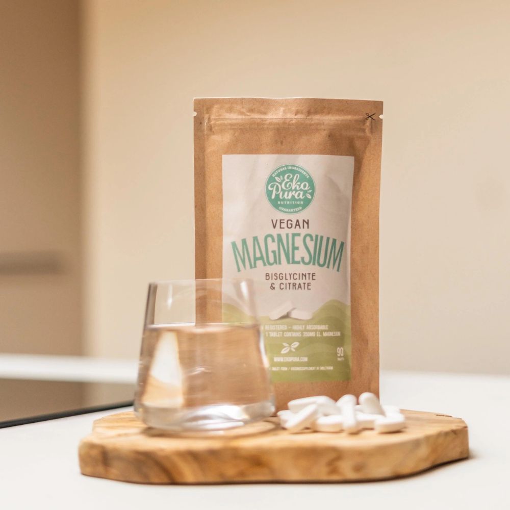 Confezione marrone con etichetta bianca. L'etichetta riporta "Vegan Magnesium". Accanto, un bicchiere d'acqua e compresse.