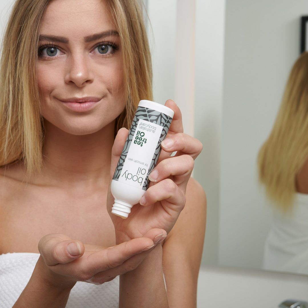 Donna tiene in mano una piccola bottiglia di olio per il corpo. Bottiglia bianca con motivi a foglie grigie. Testo: Tea Tree Oil, Australian Bodycare.