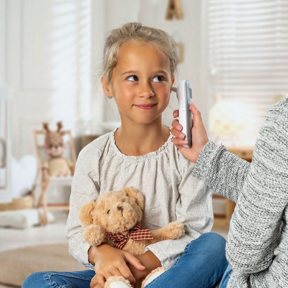 Bambina con termometro auricolare all'orecchio. Persona che tiene il termometro. Orsacchiotto sullo sfondo.