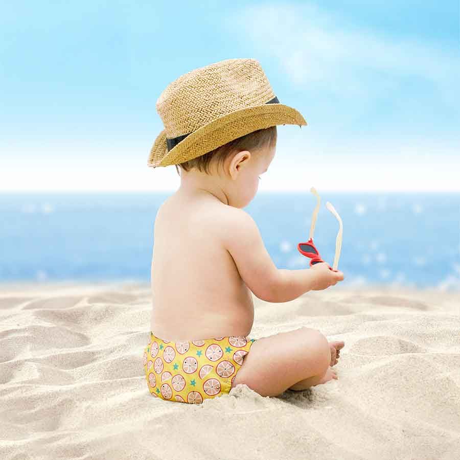 Bambino sulla spiaggia, cappello di paglia e occhiali da sole. Costumino giallo con motivi di agrumi. Mare sullo sfondo.