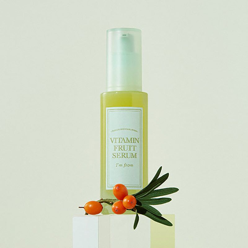 Flacone di siero con ramo di olivello spinoso. Scritta: Vitamin Fruit Serum, I'm from. Su piedistallo.