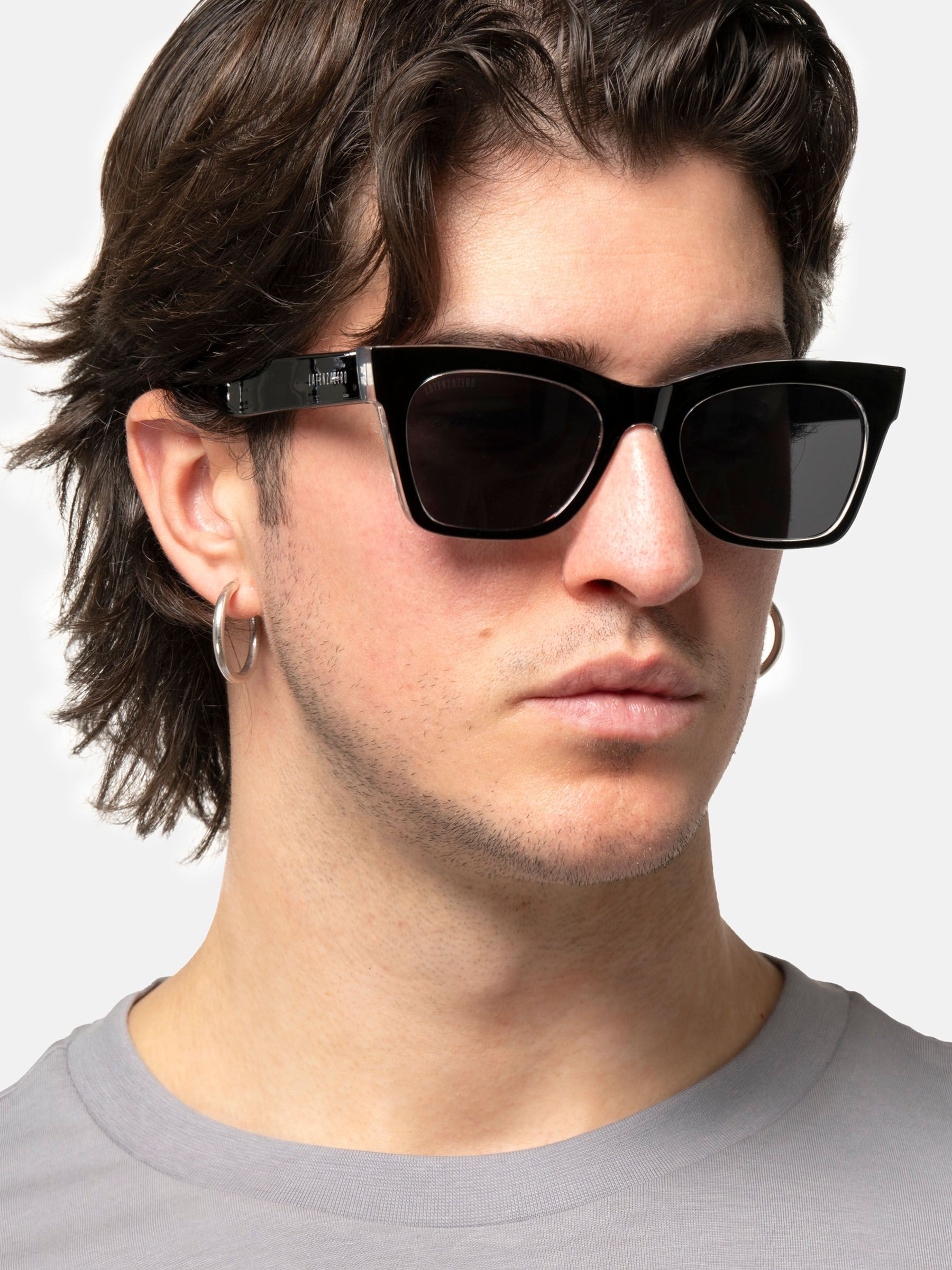 Uomo con occhiali da sole neri con lenti squadrate. Il nome del marchio è visibile sulla stanghetta.
