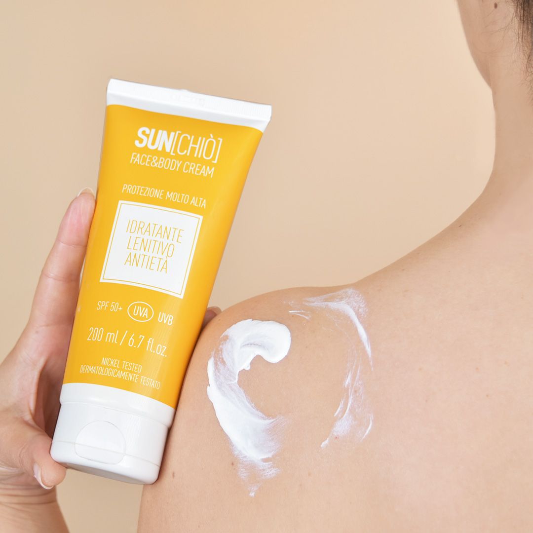 Tubo giallo con crema bianca applicata sulla spalla. Testo: SUN[CHIO], FACE&BODY CREAM, Protezione Molto Alta, Idratante Lenitivo Antietà, SPF 50+.