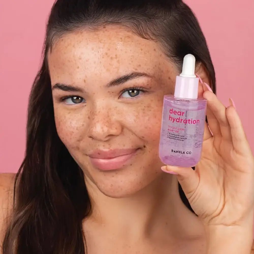 Donna tiene in mano BANILA CO Dear Hydration Crystal Glow Essence. Flacone rosa con tappo e pipetta bianchi.