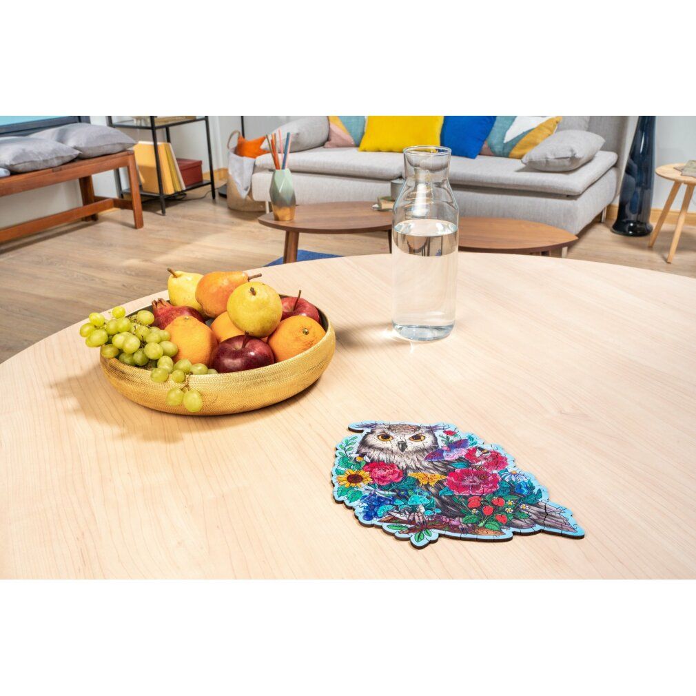 Puzzle finito con gufo e fiori su tavolo in legno. Sullo sfondo: ciotola di frutta, caraffa d'acqua.