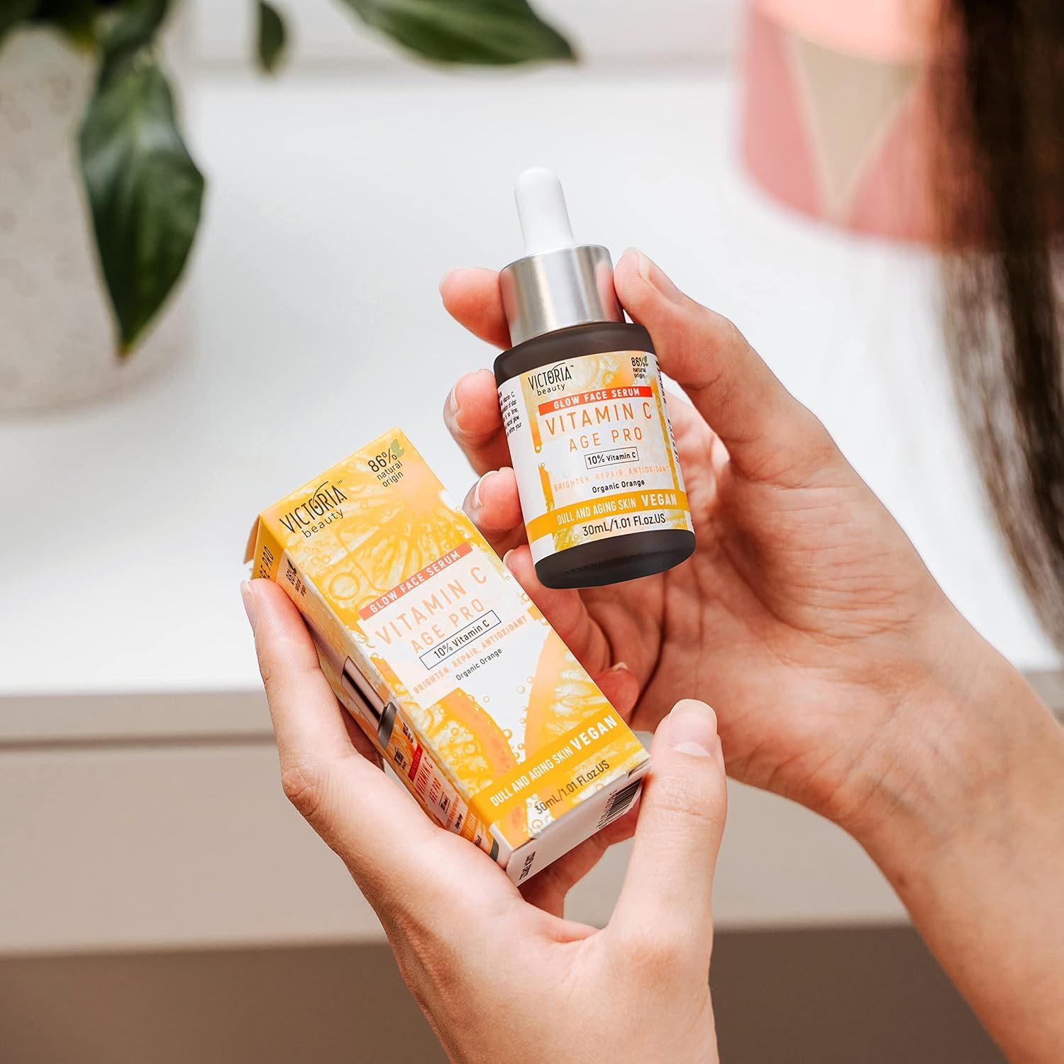 Mani che tengono flacone di siero e scatola. Scritta: Vitamin C Age Pro. Confezione gialla.