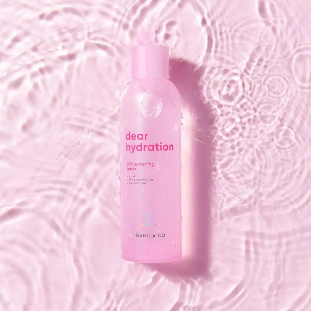 Flacone rosa "dear hydration skin softening toner" nell'acqua. Le onde creano uno sfondo rosa.