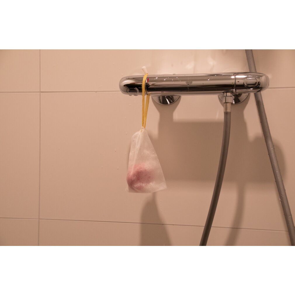 Barra di shampoo rosa in un sacchetto di rete bianco, attaccata a un rubinetto della doccia con un cordino giallo.