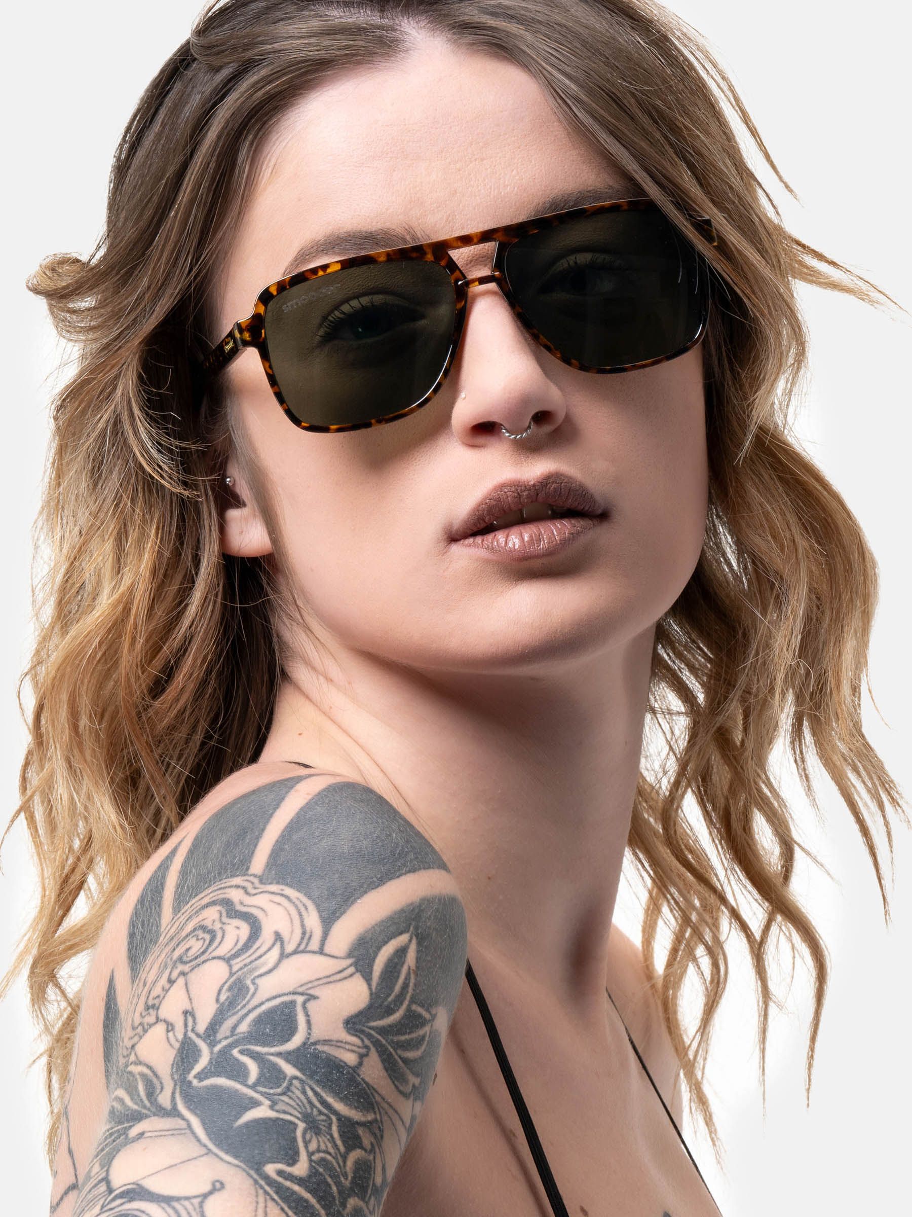 Una donna indossa occhiali da sole marroni con motivo tartarugato e lenti verdi. Gli occhiali sono di forma quadrata. La donna ha un piercing al naso.