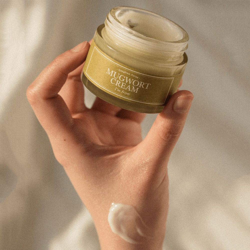 Mano che tiene un vasetto aperto. Scritta: Mugwort Cream, I'm from. Goccia di crema sulla mano.