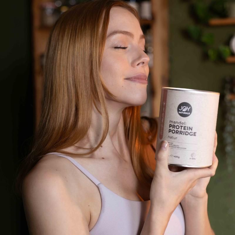 Donna tiene barattolo 'JOY NATURALS Mandel Protein Porridge Natur'. Barattolo con logo e testo. Donna con occhi chiusi.