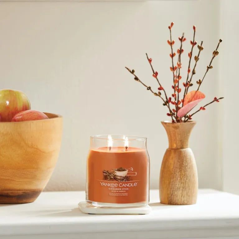 Vaso di vetro con cera arancione su un sottobicchiere bianco. Accanto, una ciotola di legno con mele e un vaso con rami.
