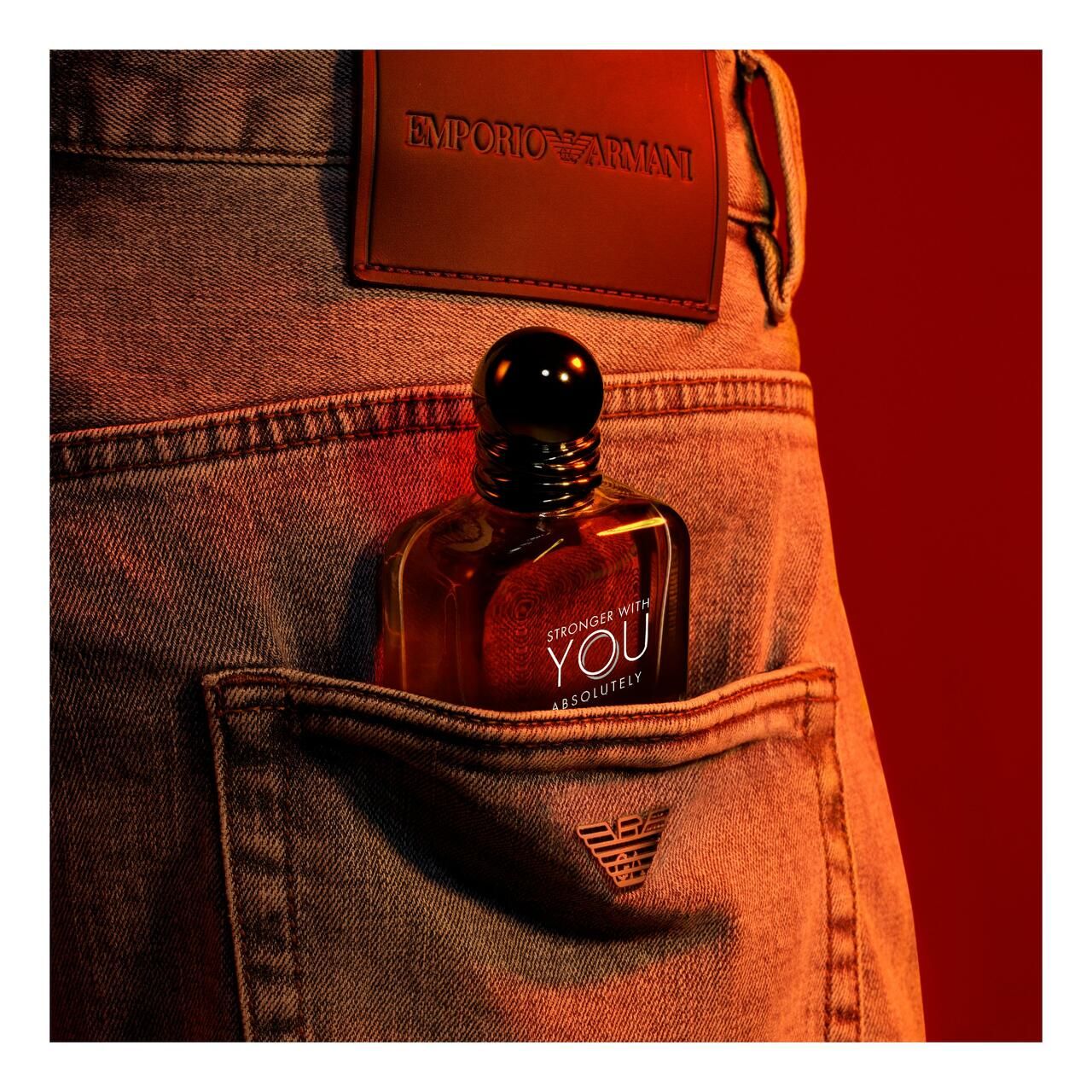 Flacone nella tasca dei jeans. Tappo nero, liquido arancione. Etichetta in pelle con logo. Sfondo rosso.