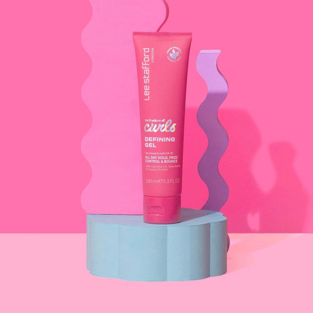 Tubetto rosa Lee Stafford For The Love Of Curls Defining Gel su un piedistallo blu, sfondo rosa.