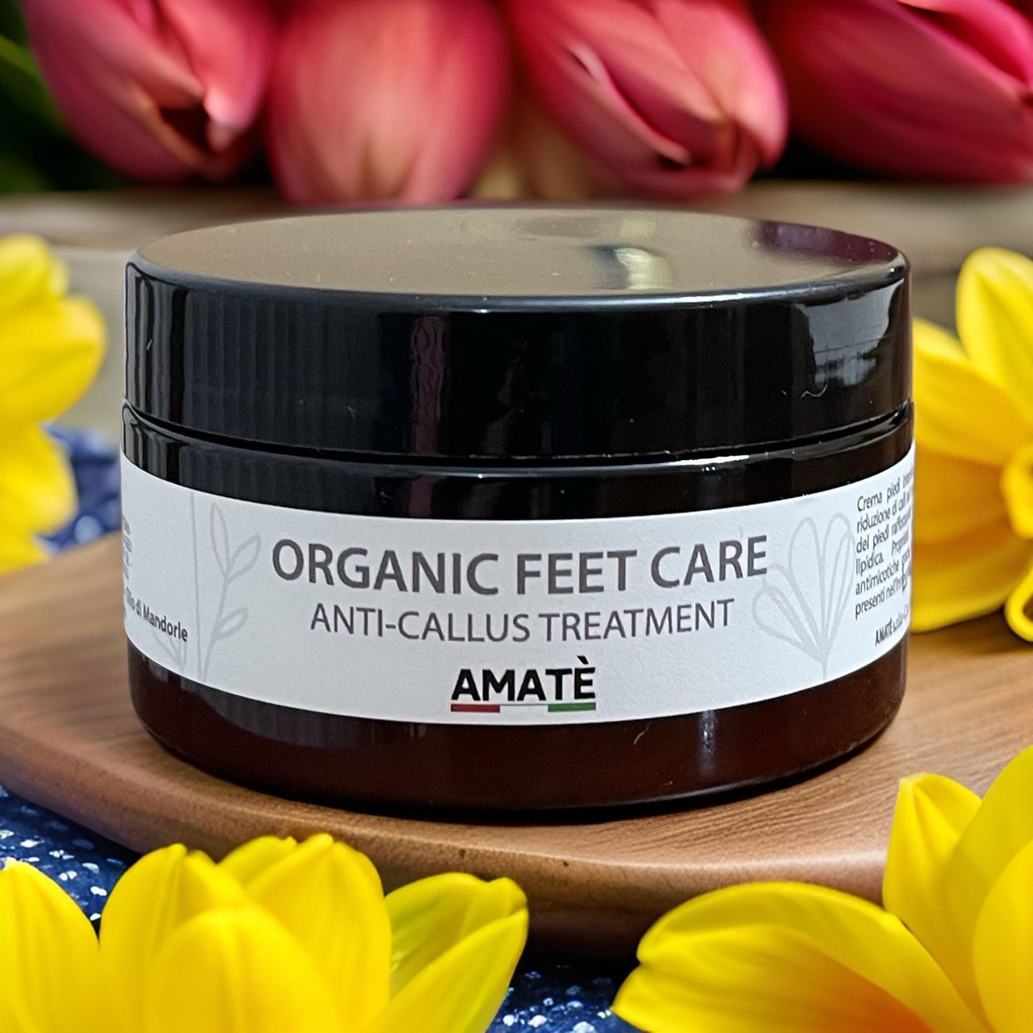 Vasetto marrone scuro con coperchio nero. Scritta: ORGANIC FEET CARE ANTI-CALLUS TREATMENT. Marchio: AMATÈ. Su tagliere.
