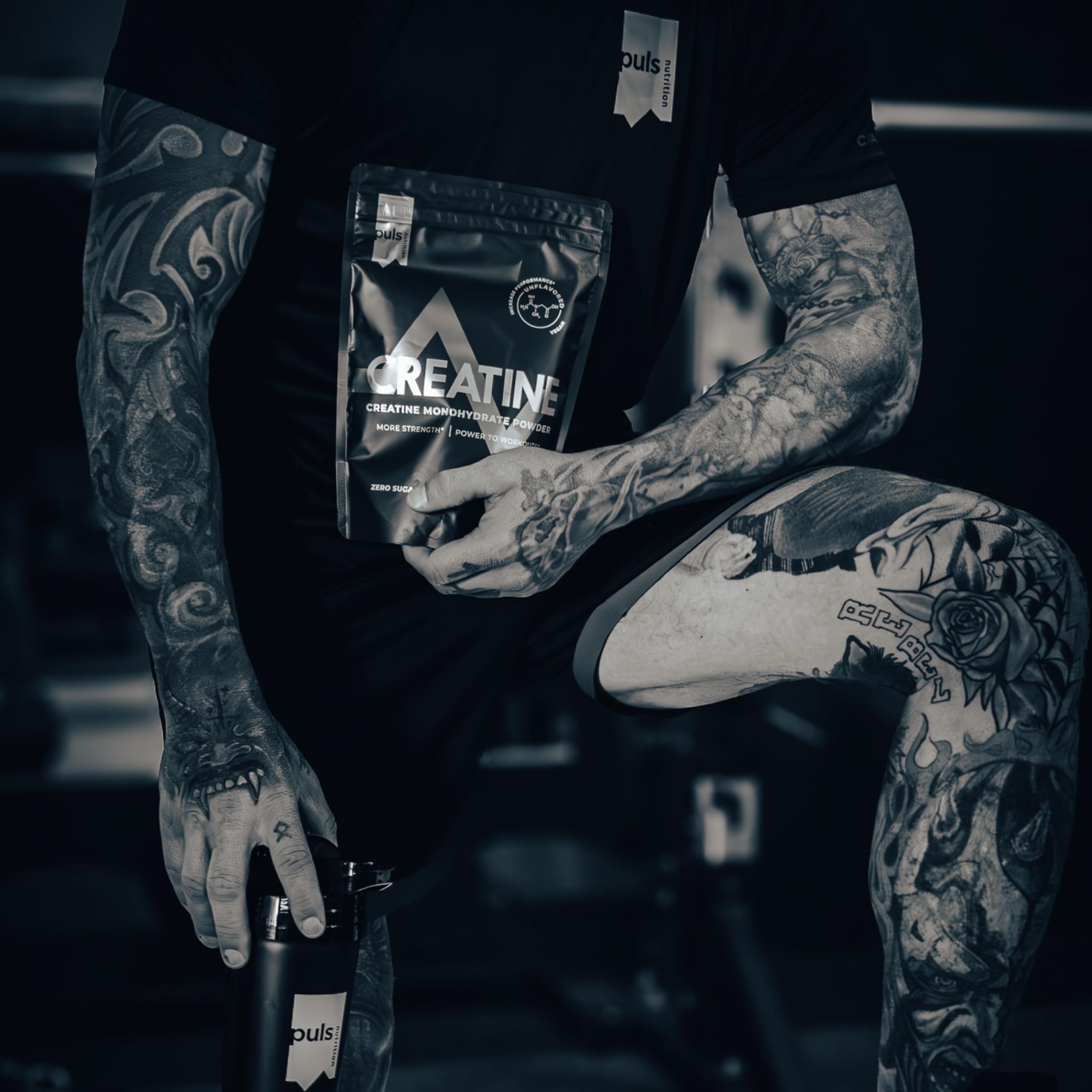 Uomo tatuato tiene un pacco di polvere di creatina e una bottiglia. Marchio: Puls Nutrition.