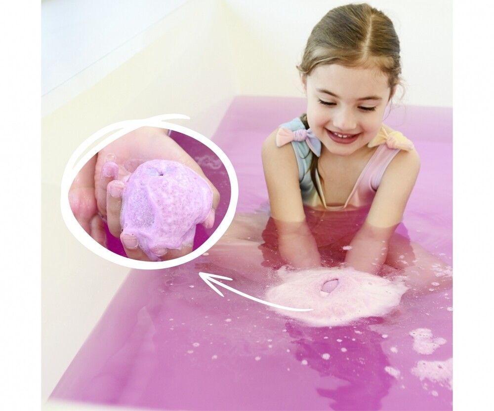 Bambina nella vasca da bagno con acqua rosa. In mano una bomba da bagno rosa che si scioglie. Una freccia indica la bomba da bagno.