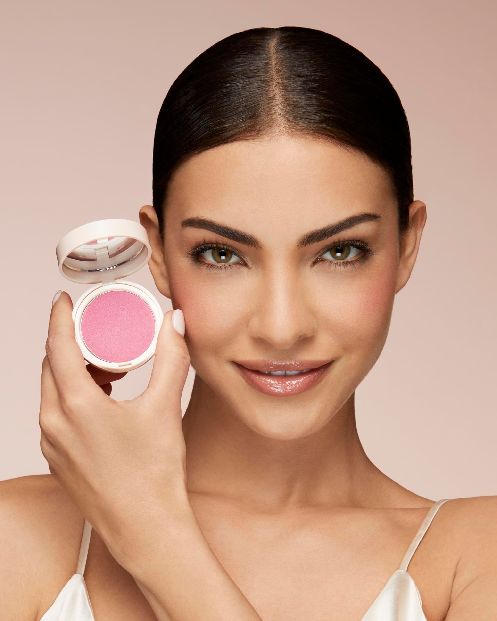 Donna tiene una polvere blush davanti alla guancia. La polvere è rosa. Sorride.