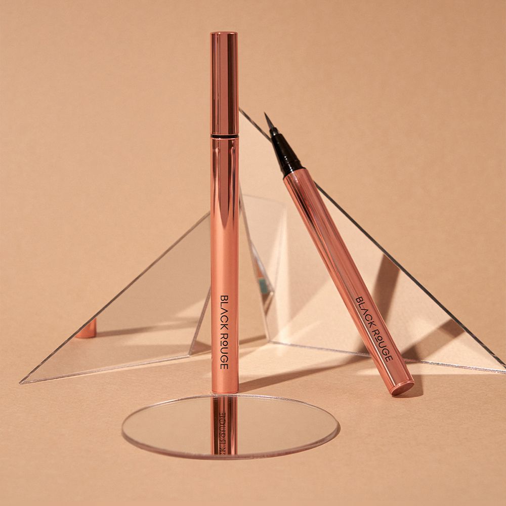 Due penne eyeliner, una in piedi, una sdraiata. Entrambe color oro rosa. Scritta: BLACK ROUGE. Specchi sullo sfondo.