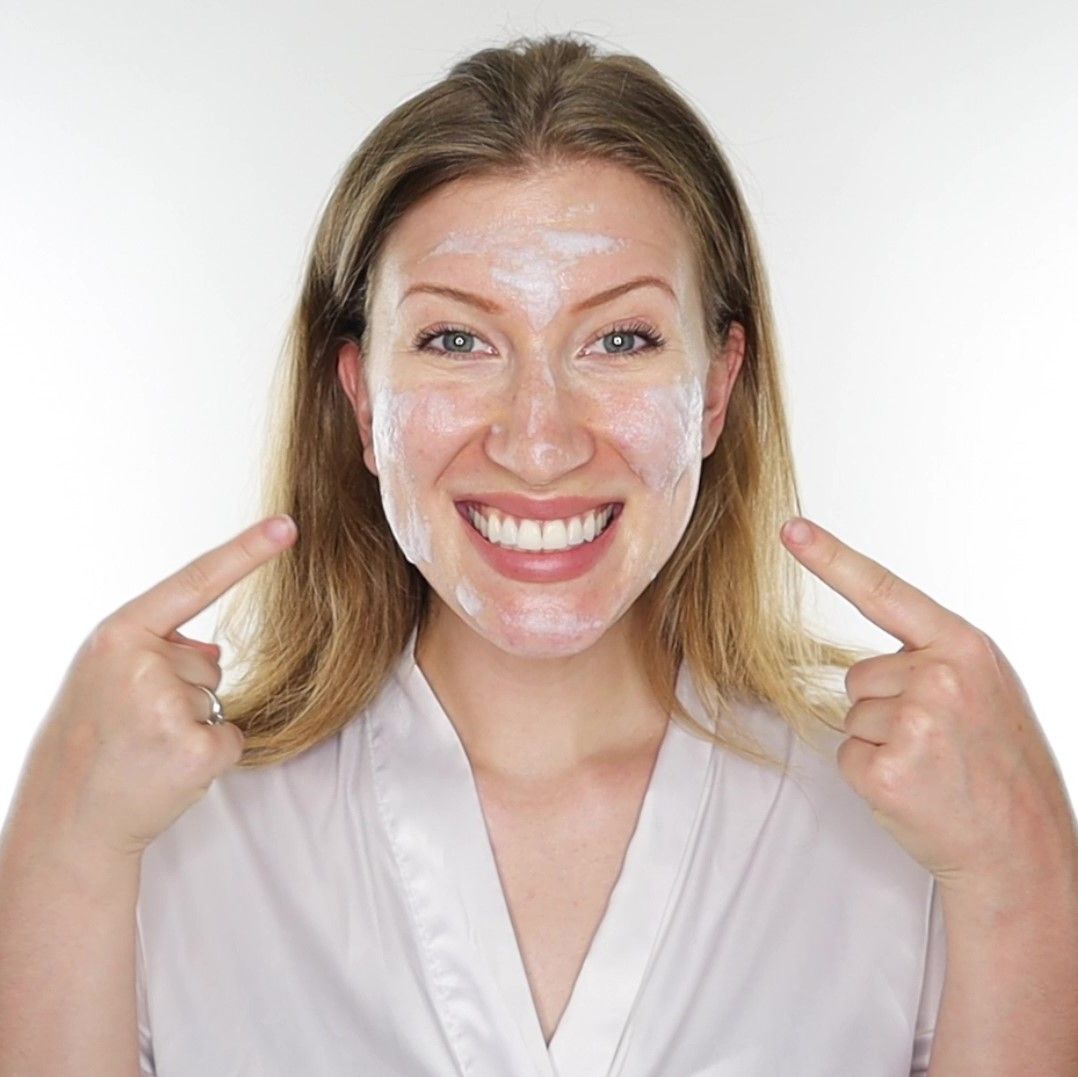 Donna con crema bianca sul viso. Indica il suo viso con le dita. Indossa un accappatoio bianco.