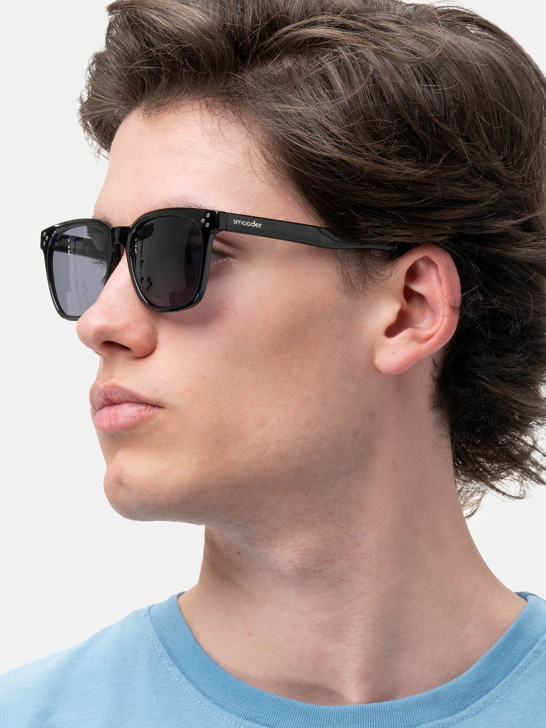Uomo con occhiali da sole neri. Marchio "Smooder" sulla stanghetta. Quattro rivetti agli angoli della montatura.