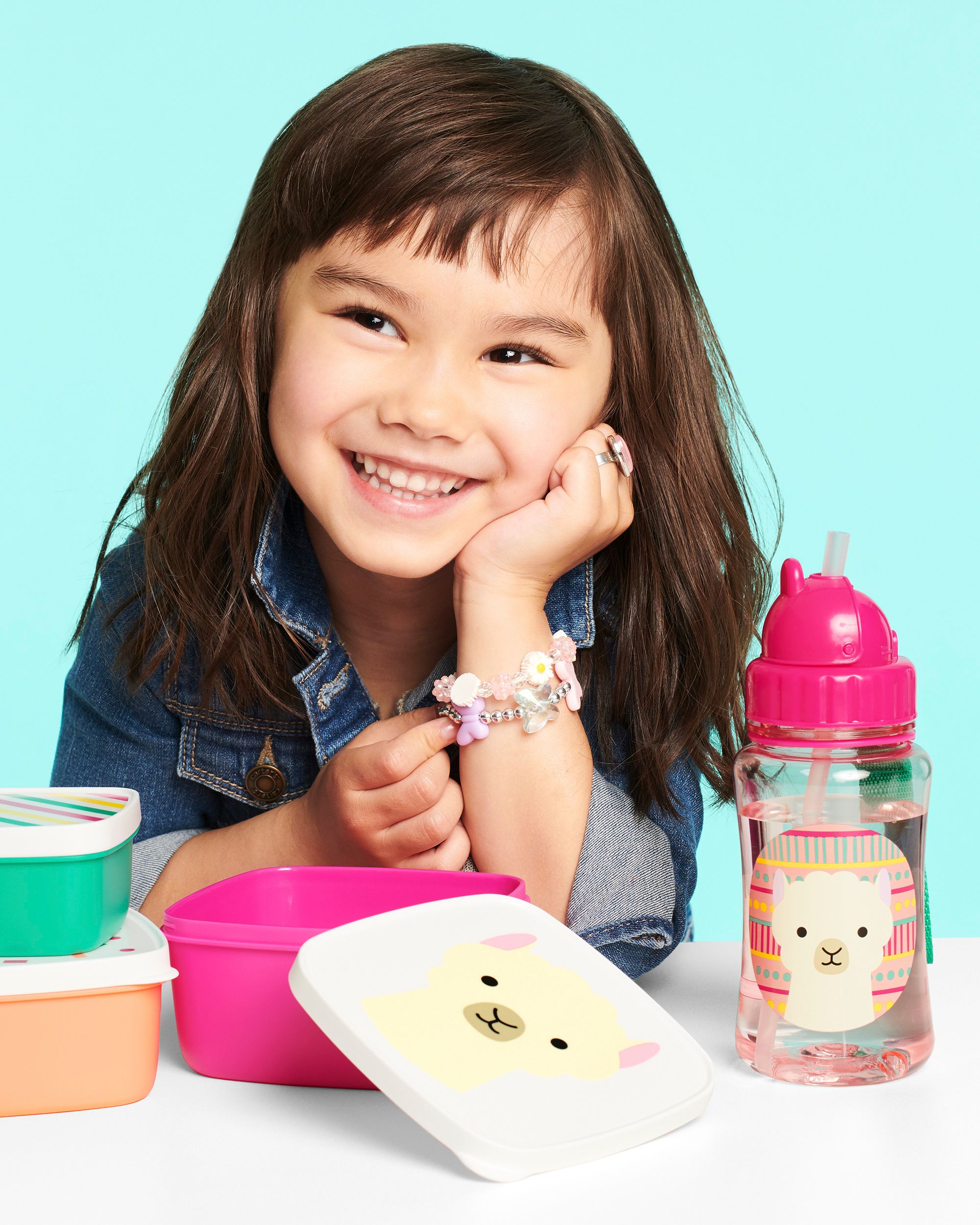 Bambina con borraccia e portapranzo. Borraccia rosa con motivo lama. Portapranzo di diversi colori. La bambina indossa una camicia di jeans.