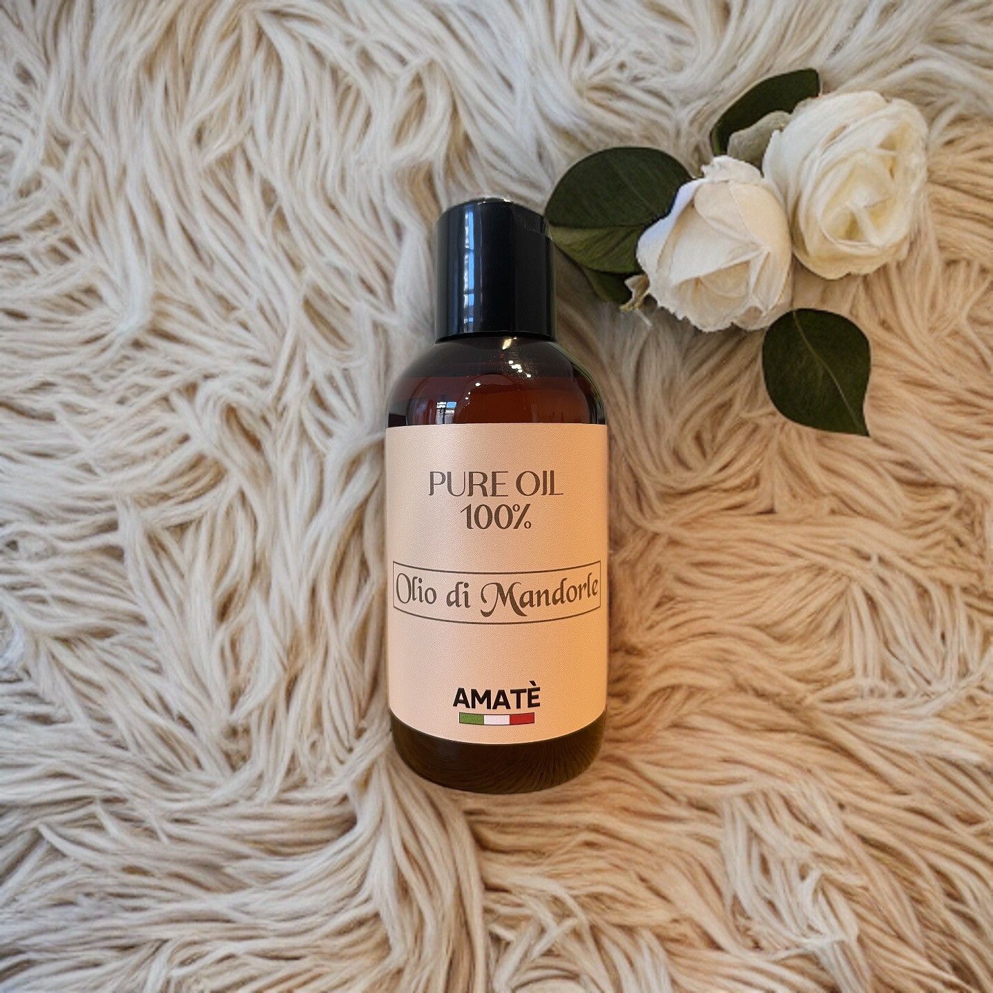 Flacone marrone con tappo nero su pelliccia bianca. Etichetta "PURE OIL 100%" e "Olio di Mandorle". Rose bianche.