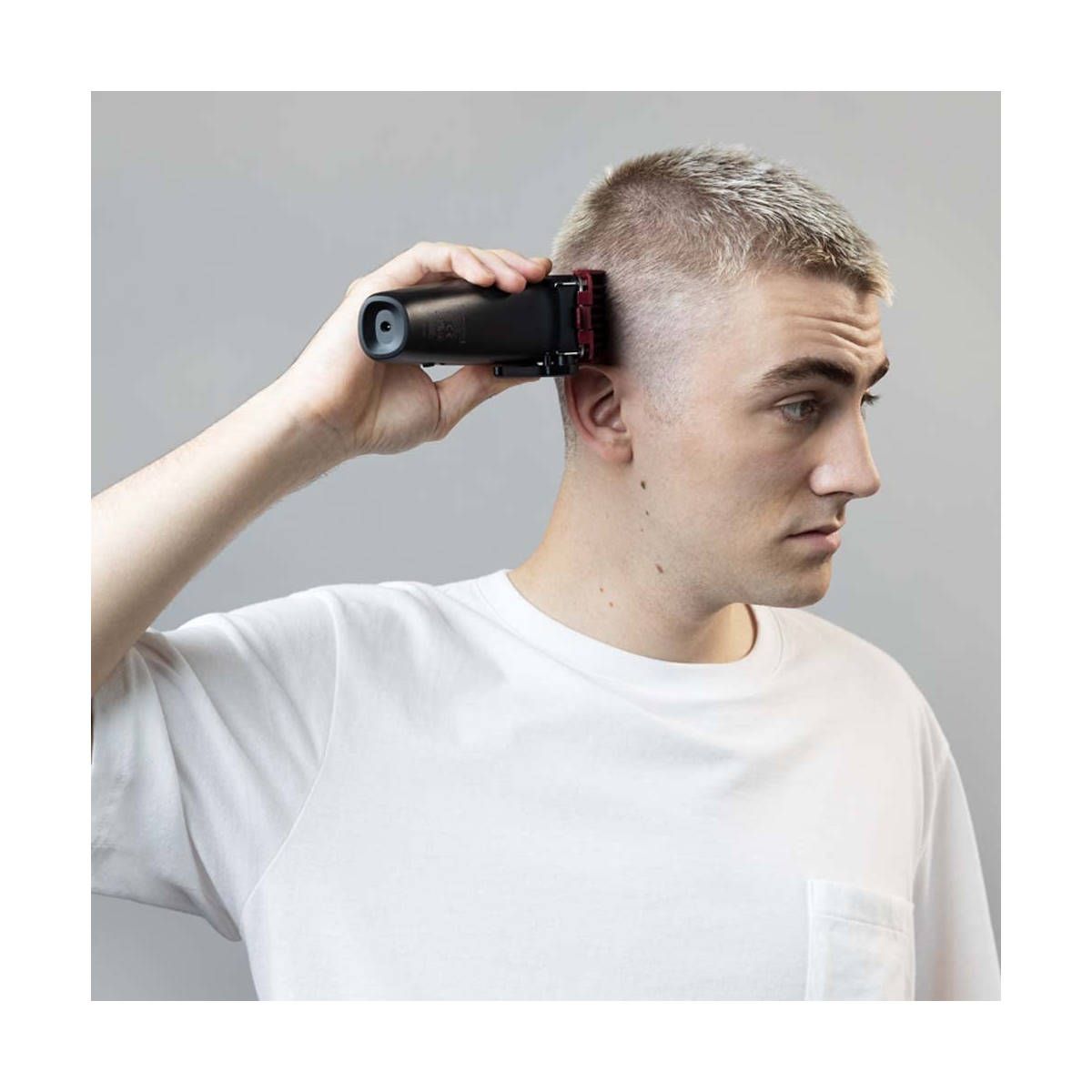 Uomo che si taglia i capelli con un tagliacapelli nero. Pettine rosso. Sfondo grigio.